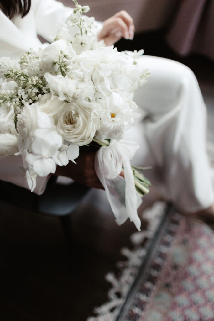 Bride holding a white floral bouquet while seated, with soft focus on flowers and wedding trousers.