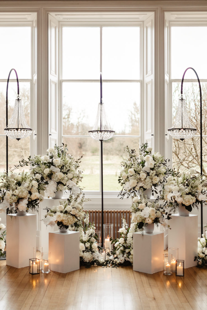 Symmetrical indoor wedding ceremony floral arrangement with white flowers, modern plinths, glass candles, and hanging chandeliers.
