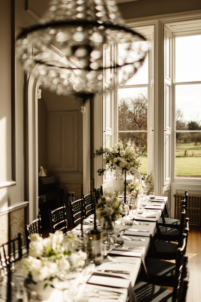 Elegant wedding table setting by a large window with natural light, white floral centerpieces, and a chandelier in soft focus.