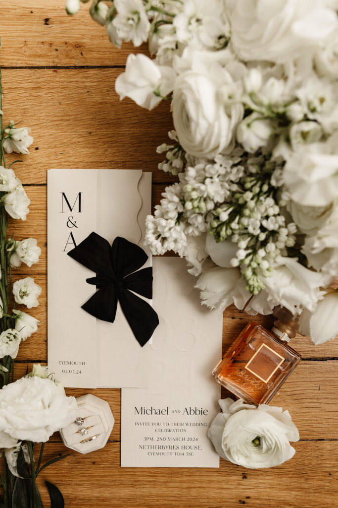 Flat lay of wedding stationery with black ribbon, white flowers, and Chanel perfume bottle on wooden surface.