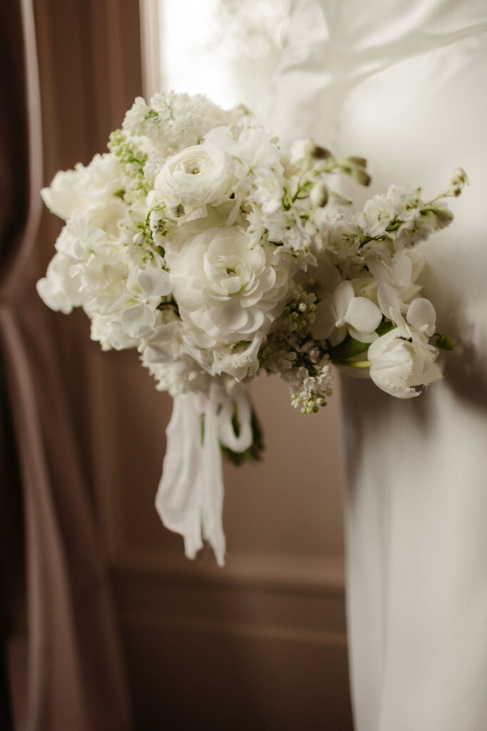 Close-up of white wedding bouquet featuring ranunculus, orchids, tulips and lily of the valley, held by bride at Netherbyres House.