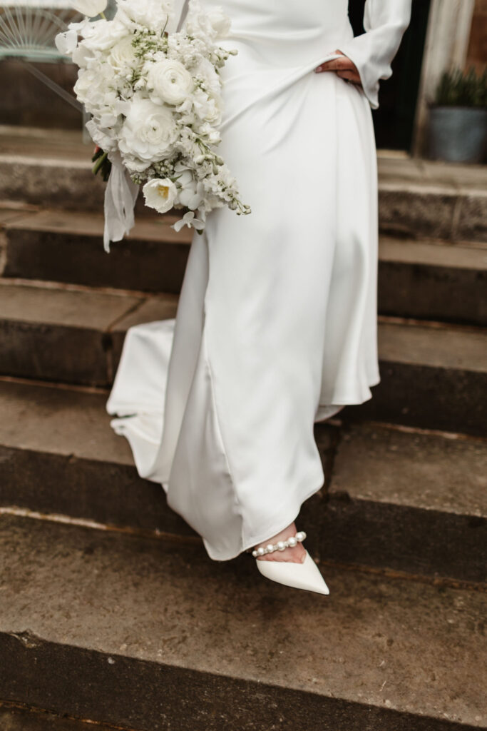 Bride descending stone steps in white satin gown, holding white bouquet and wearing pearl-detailed heels.