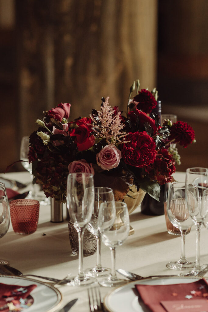Close-up of rich red and blush wedding flower centrepiece with roses and seasonal blooms on a candlelit reception table at Mount Stuart, Rothesay.